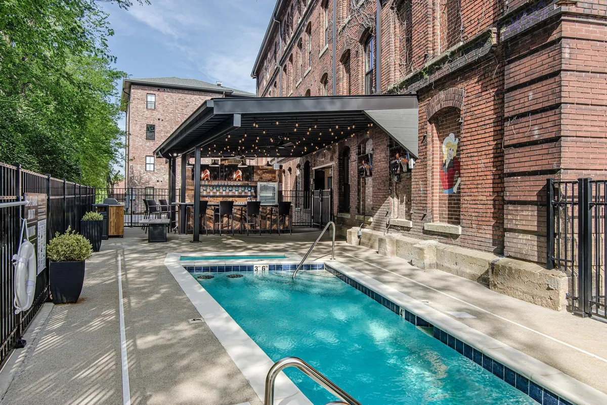 This image captures a lively outdoor pool area with a sparkling swimming pool, surrounded by a covered seating area, exposed brick walls, and string lights overhead. People are shown swimming, socializing, and relaxing in the sun, creating a vibrant atmosphere ideal for group gatherings and poolside fun.