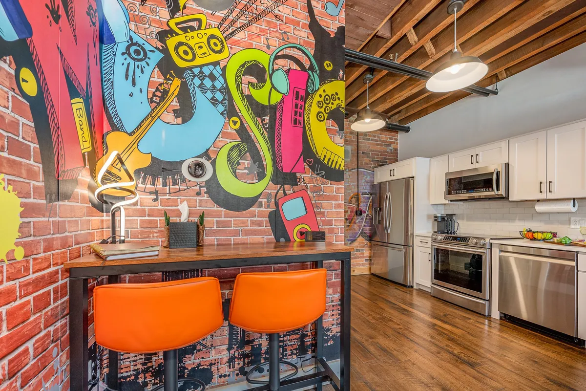 A colorful graffiti wall serves as the backdrop to orange bar stools and stainless steel appliances. The playful, creative mood is enhanced by the artistic decor and modern furnishings, suggesting a space perfect for both work and leisure.