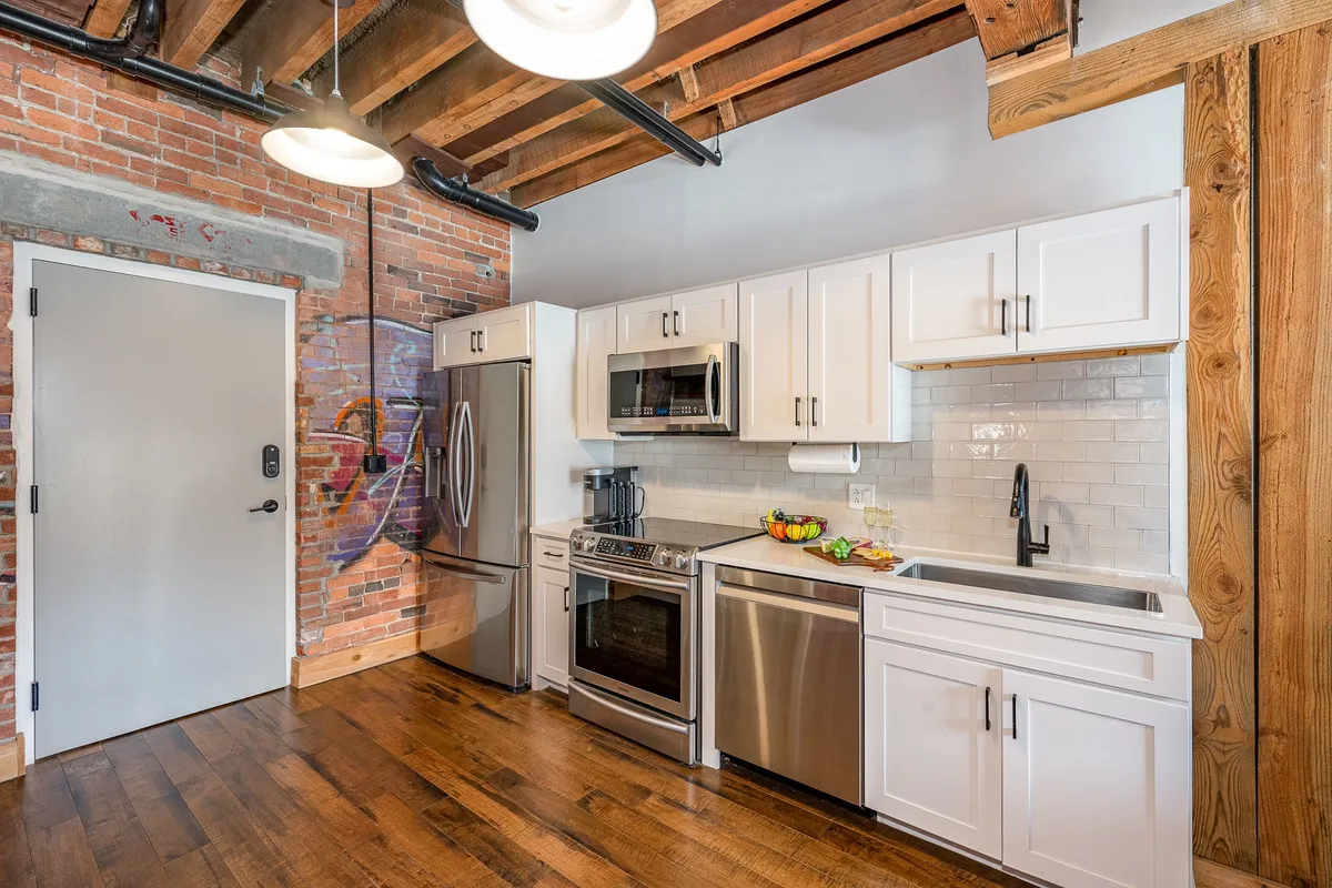 A vibrant, contemporary kitchen featuring stainless steel appliances, white cabinetry, an exposed brick wall, wooden beams overhead, and an artistic mural. The layout showcases a kitchen island and open space ideal for cooking and socializing. The five-burner stove is visible among the appliances, and the kitchen feels bright and energetic.