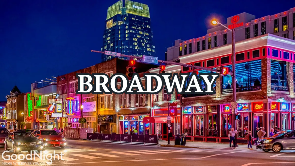 A vibrant shot of Broadway at night, featuring illuminated neon signs, a buzzing street filled with people, cars, and high-rise buildings in the background. The energetic atmosphere showcases the heart of Nashville’s famous live music and nightlife district.