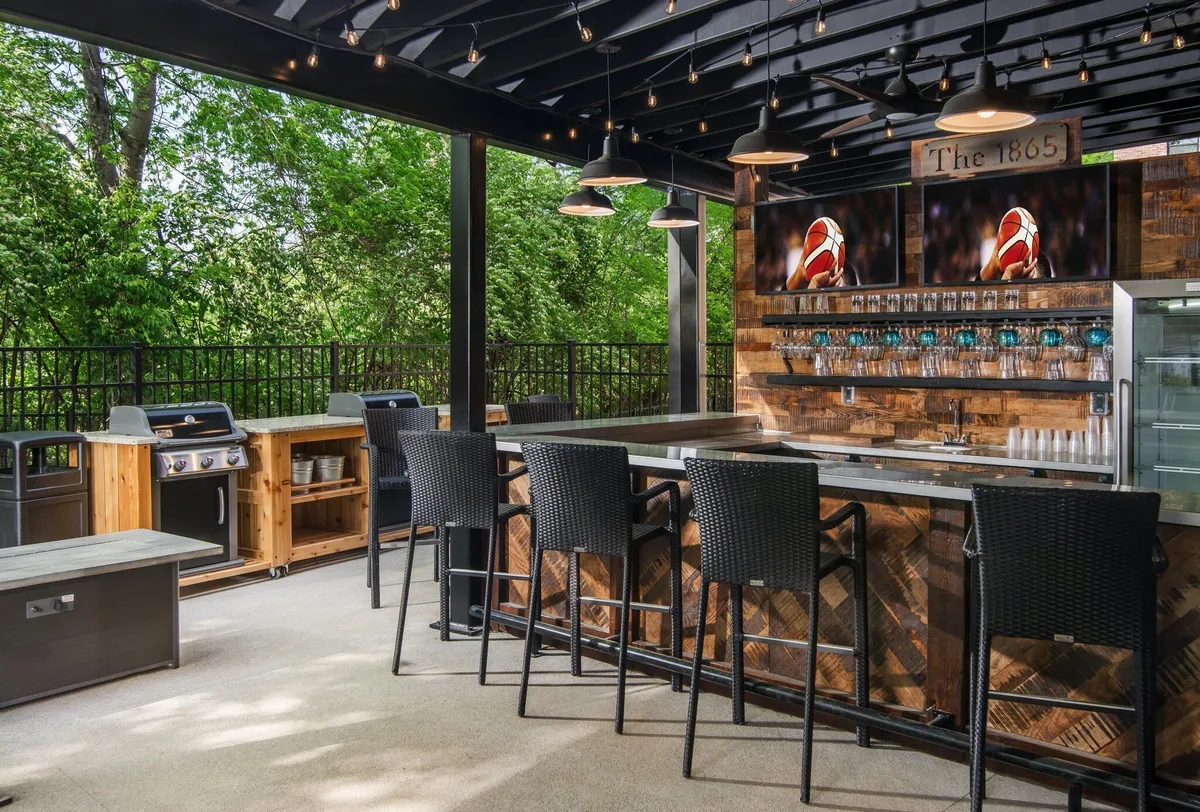A bustling exterior lounge space featuring a modern bar counter, outdoor grill stations, TV screens, hanging lights, and sleek wood paneling. The setting is lively, with groups of people entertaining, barbecuing, and enjoying drinks while watching sports, evoking the spirit of summer cocktail parties and communal cookouts.