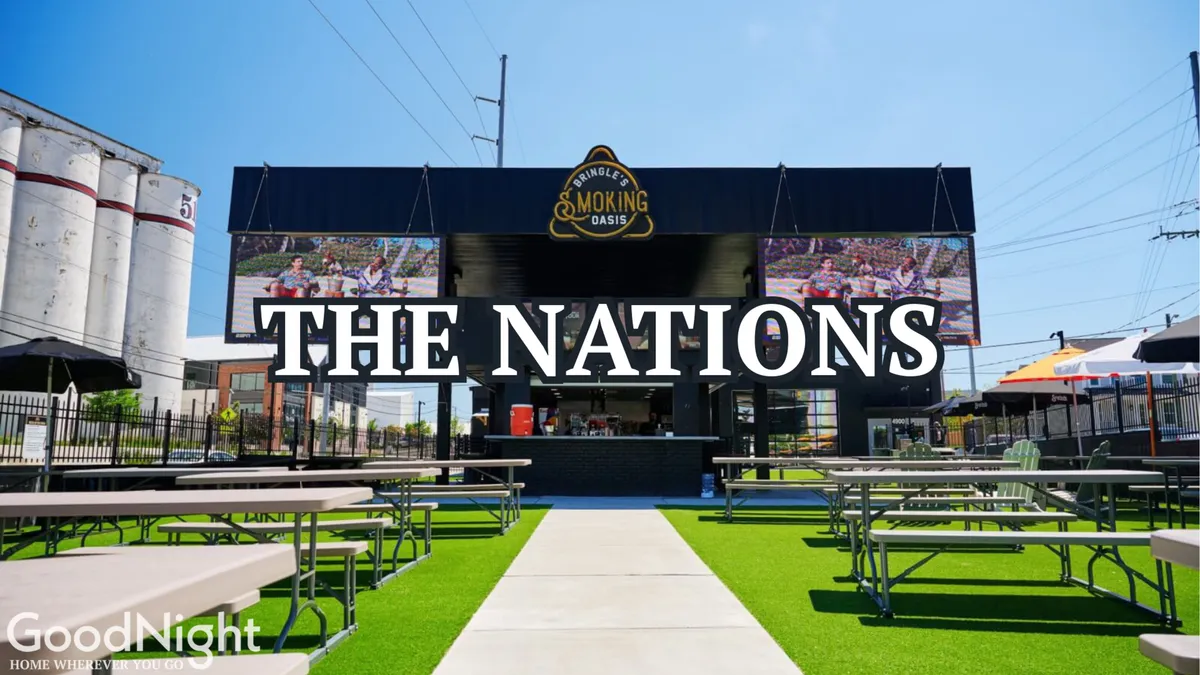 A lively outdoor seating area in front of a building with prominent signage and large screens displaying events, surrounded by groups of people enjoying food and drinks together in a vibrant, communal atmosphere. The open-air environment is perfect for social gatherings and watching live sports or communal TV events under the sky.