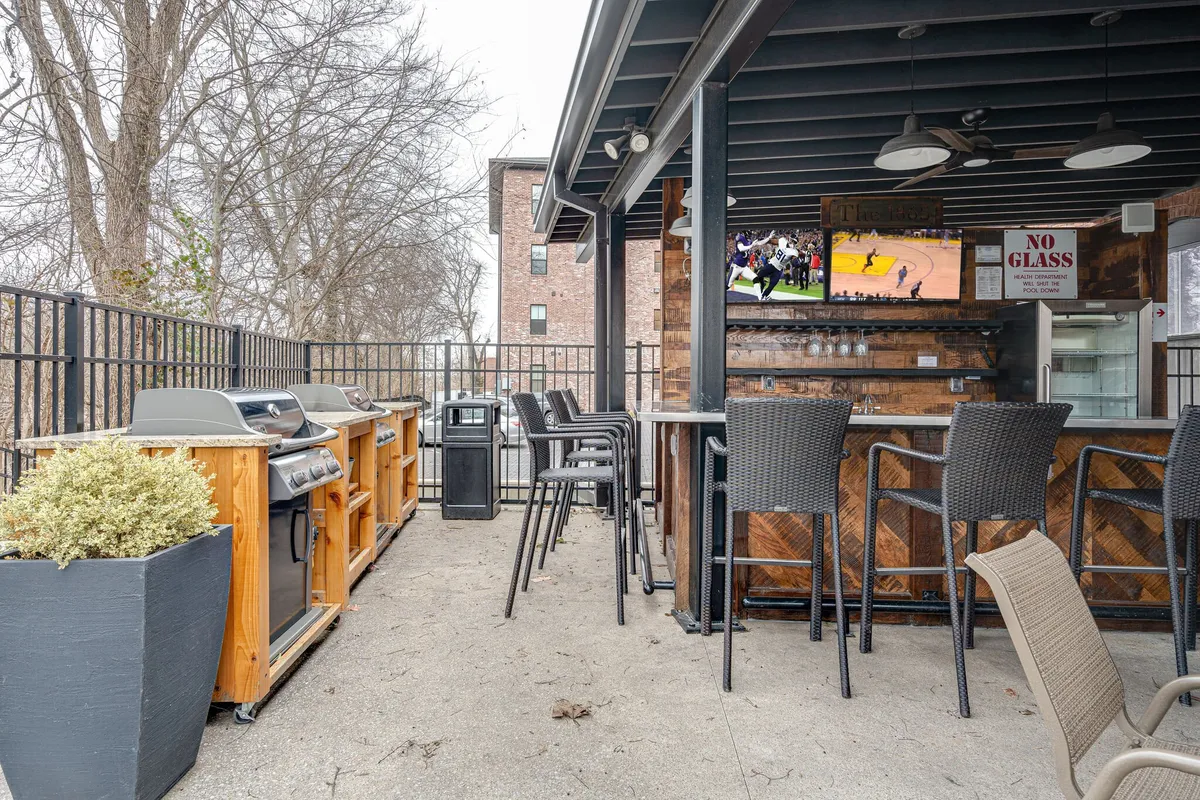 A stylish exterior patio area with grills, a bar, a television screen, and plenty of comfortable seating, inviting residents to gather outdoors for meals, drinks, and entertainment. The space buzzes with relaxed energy and sociable vibes.