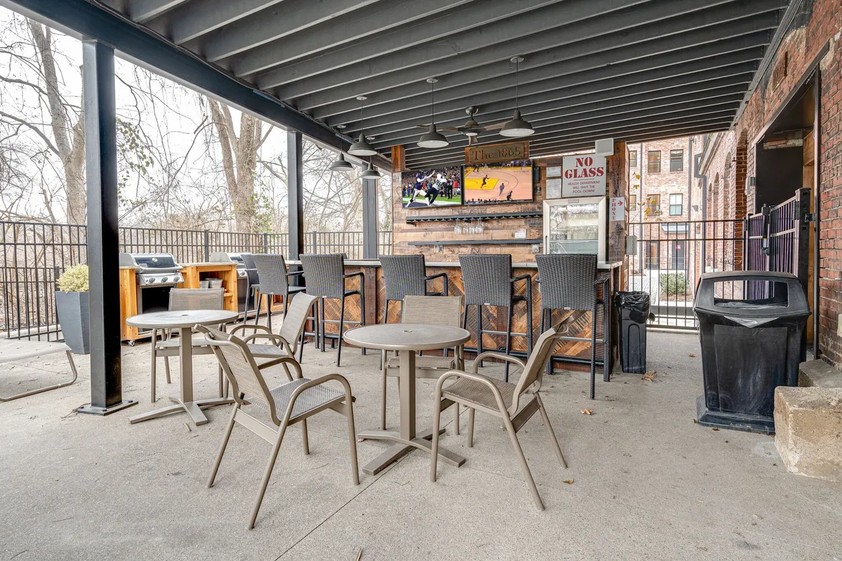 An inviting outdoor seating area featuring a large television screen, grill area, and bar counter, with groups of people socializing and relaxing in the sun. The setup encourages both leisure and community, reflecting the lively spirit of shared amenities.