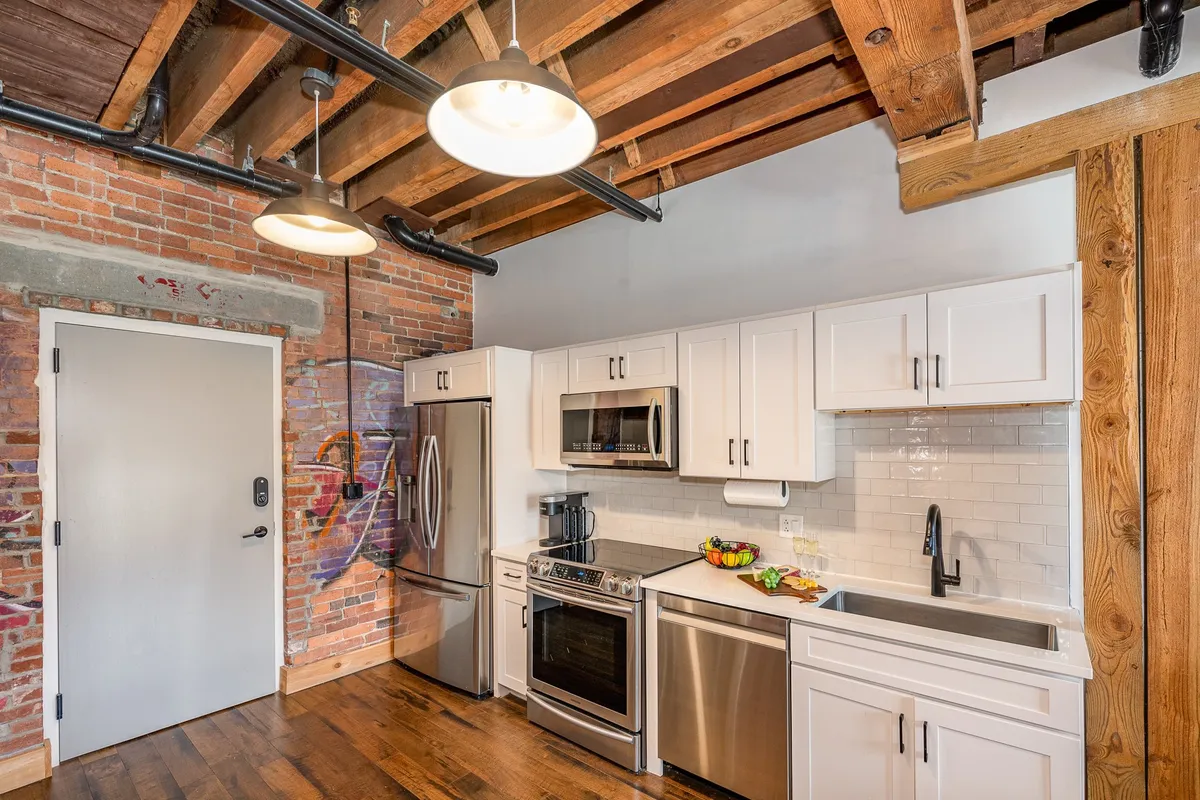 An open kitchen with an exposed brick wall, stainless steel appliances, open shelving, and warm wooden beams. The space feels cozy yet contemporary, with functional features for both cooking and entertaining. The layout allows for easy movement between cooking and dining areas.