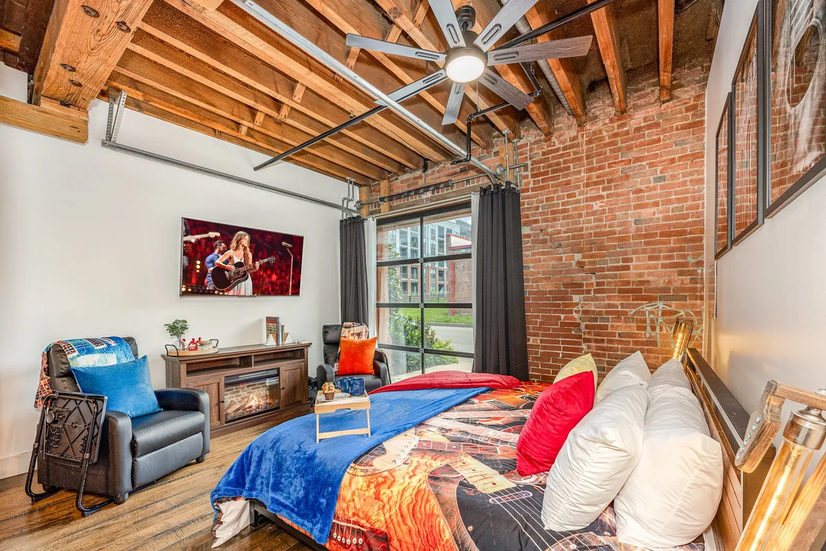 This image showcases a bed with vibrant bedding, a large window with a view, a television displaying a performance, and recliner chairs. The scene feels inviting and creative, with natural light streaming into the room.